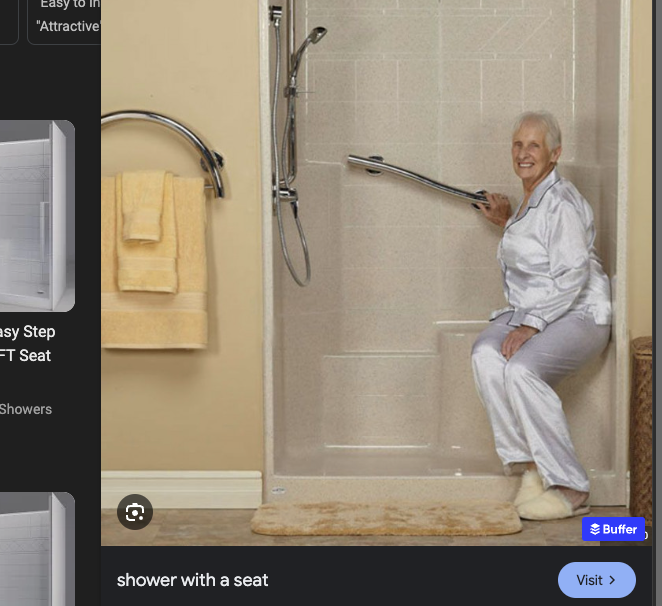 Picture of a woman with short gray hair sitting on a seat in the back of a shower. She has on slippers and silk pajamas. She has one hand on a grab bar. There is a hand held shower head that's adjustable in the front of the shower.